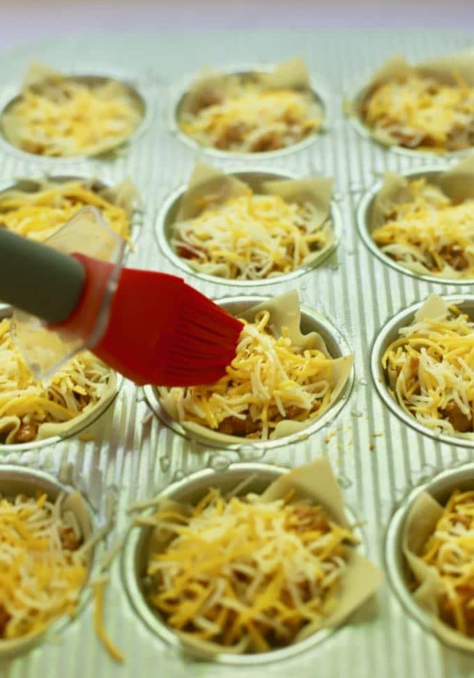 Using a pastry brush to brush on vegetable oil to wonton wrappers in a muffin pan full of taco meat. 