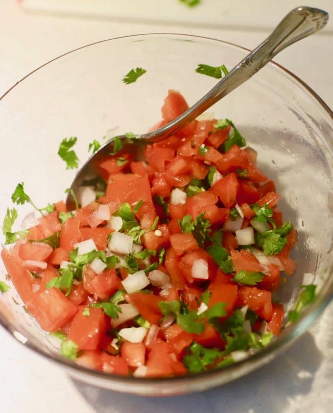A clear glass mixing bowl full chopped tomatoes, cilantro, and onions.