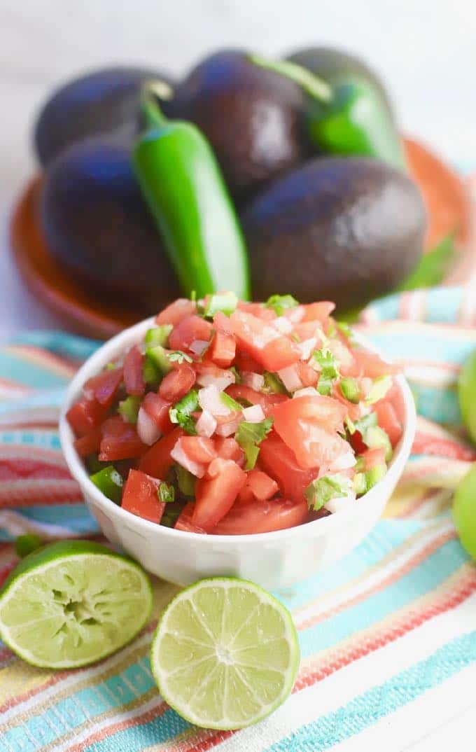 A white bowl full of pico de gallo on a brightly colored dish towel with avocados and jalapeños in the background.