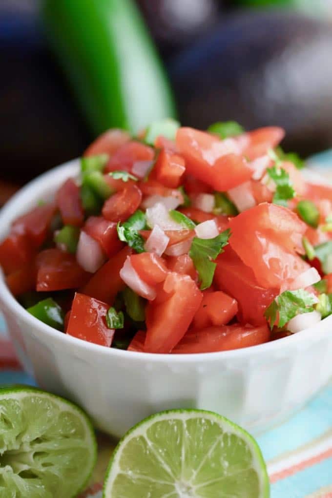 A mixture of chopped tomatoes, onions, and cilantro in a white bowl with cut limes in front.