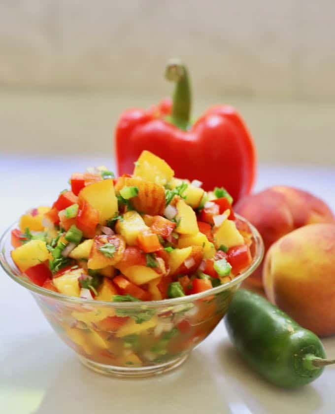 A clear glass bowl of Easy Southern Fresh Peach Salsa with a red bell pepper, peaches and a jalapeno pepper next to it.