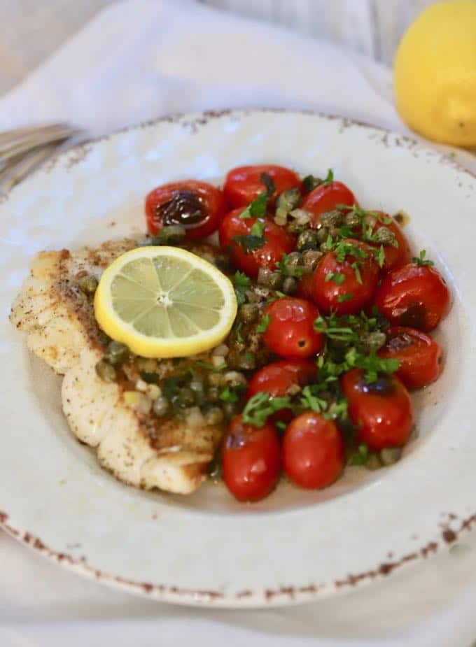 Pan fried fish with blistered tomatoes and a lemon parsley butter sauce garnished with a lemon slice on a white plate