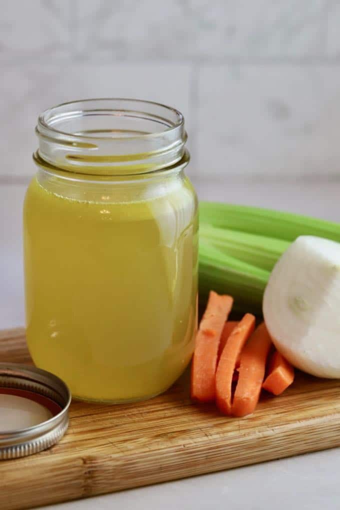 A jar of homemade chicken stock with carrots and celery on a cutting board.