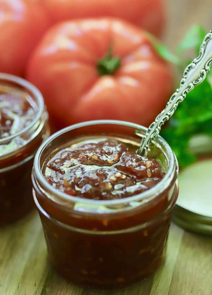 An open jar of tomato jam with fresh tomatoes in the background.