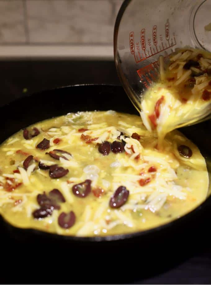 Pouring egg, shredded cheese, olives and sun-dried tomatoes into a pan.