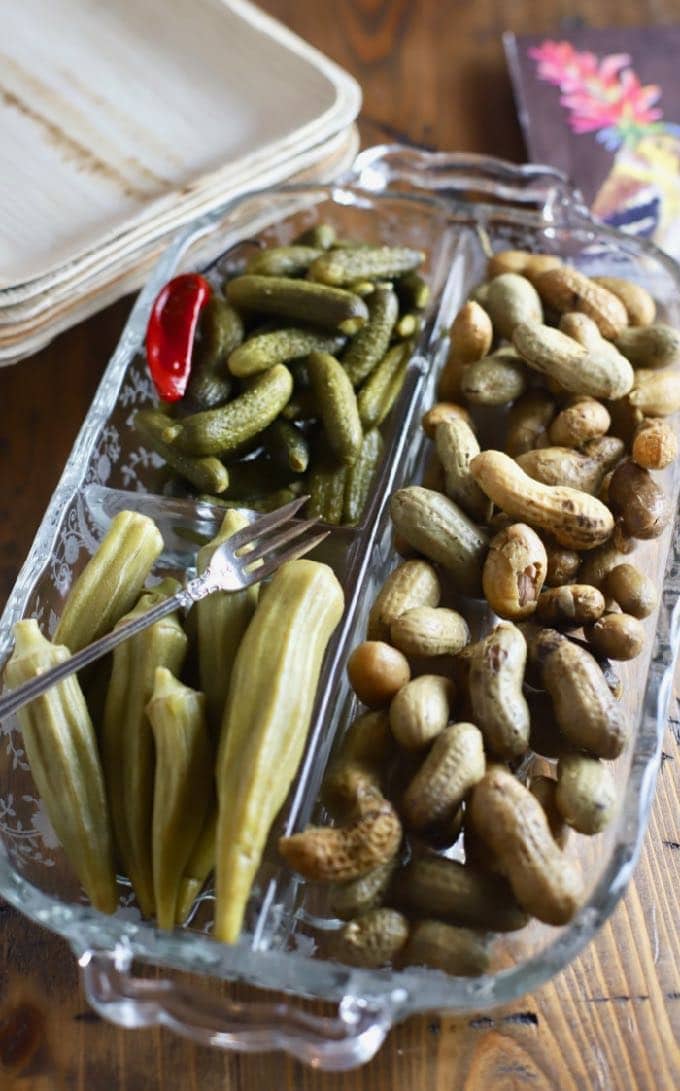 A glass dish with pickled okra, boiled peanuts and gherkins.