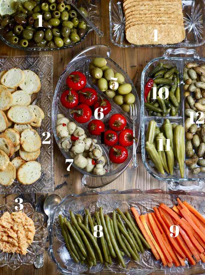 Seven glass trays on a board filled with relish tray items like olives and pickles.