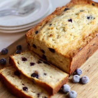 A loaf of banana bread with blueberries on a cutting board with two slices cut.