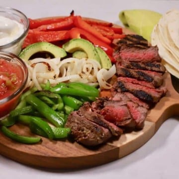 Fajita ingredients on a cutting board including steak, peppers, onions, and tortillas.