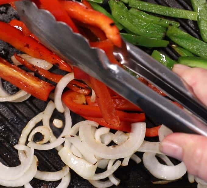 Red and green bell pepper slices and sliced onions cooking on a grill pan on the stove.