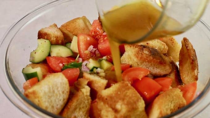 Adding dressing to panzanella salad in a clear glass bowl.