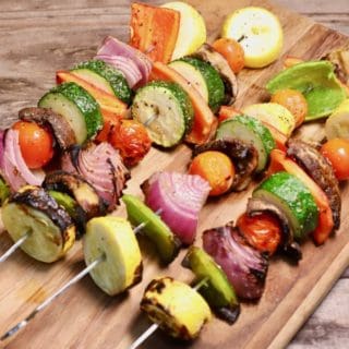 Four grilled veggie skewers on a wooden cutting board.