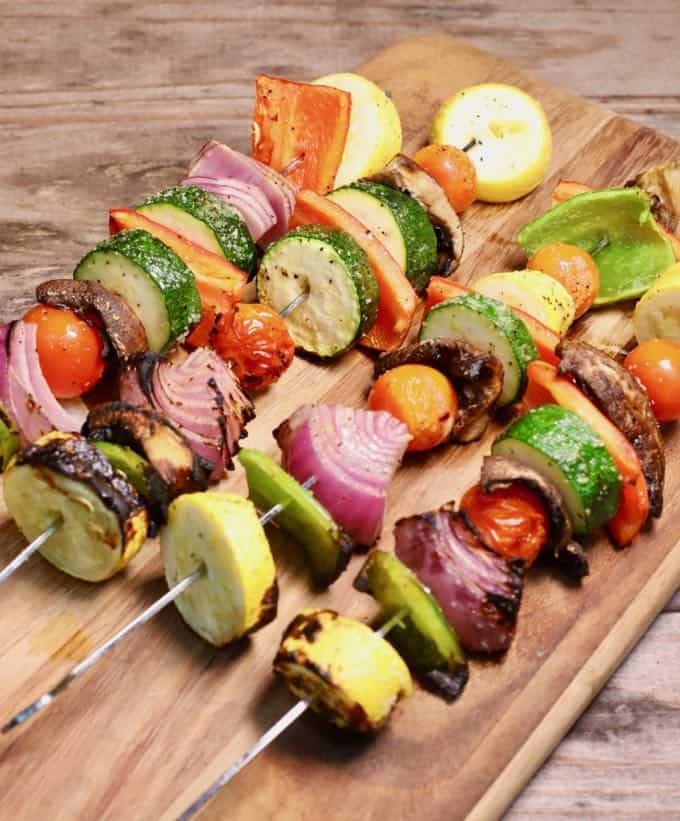 Four grilled veggie skewers on a wooden cutting board.