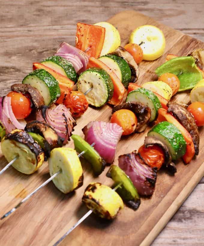 Four grilled veggie skewers on a wooden cutting board. 