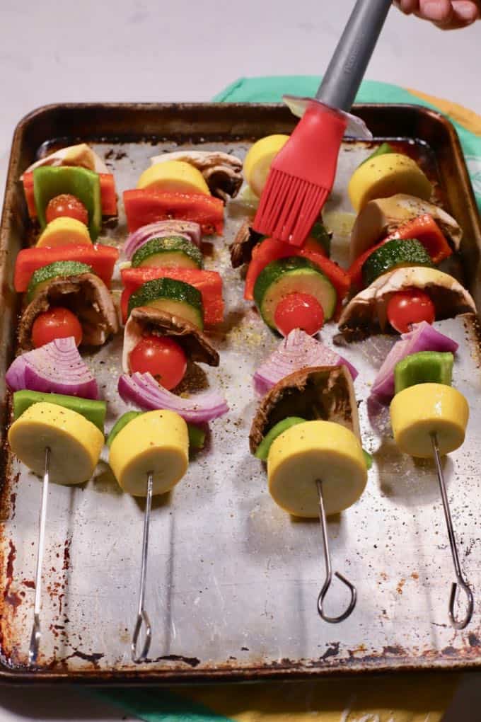 Using a pastry brush to all olive oil to four vegetable kabobs on a baking sheet.