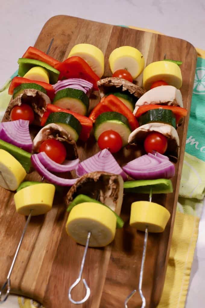 Four vegetable kabobs on metal skewers on a wooden cutting board