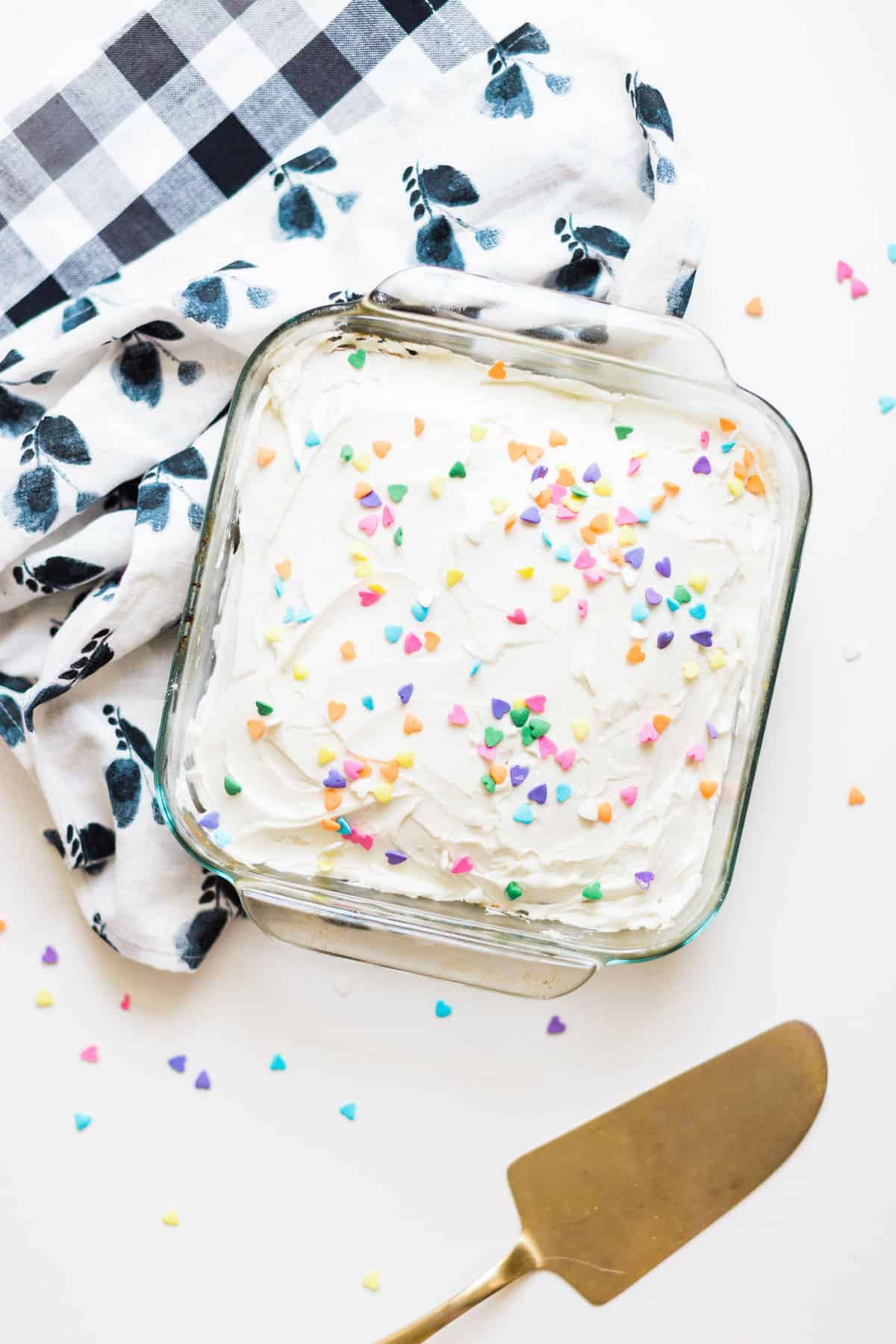 A glass baking dish with vanilla sheet cake topped with icing and sprinkles.