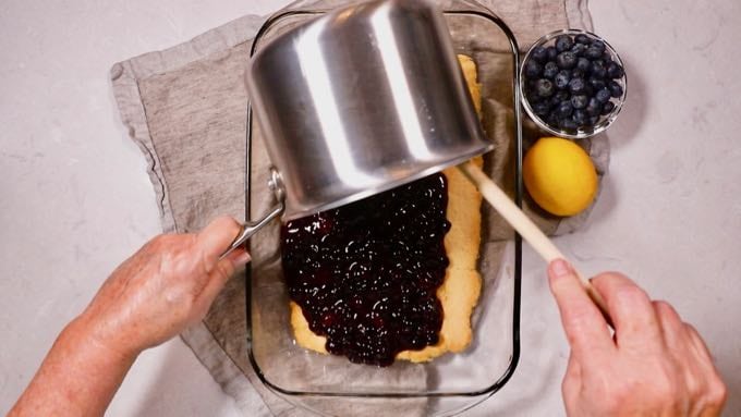 Pouring a cooked blueberry filling over a crust in a baking dish. 