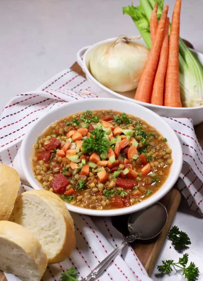 A white bowl full of lentil soup with carrots, celery and an onion in a bowl behind it.