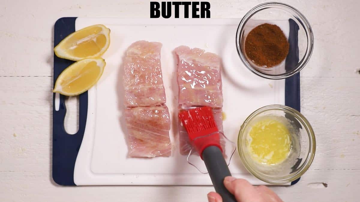 Brushing butter on two fish fillets on a cutting board.