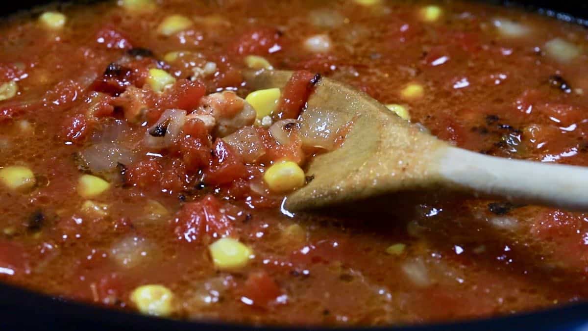A wooden spoon stirring soup ingredients including canned tomatoes and corn.