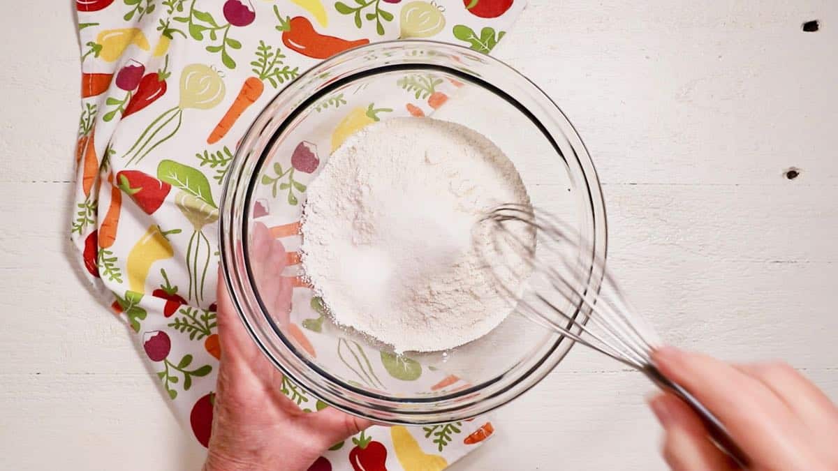 Using a whisk to combine flour, cornmeal and sugar in a clear glass bowl.