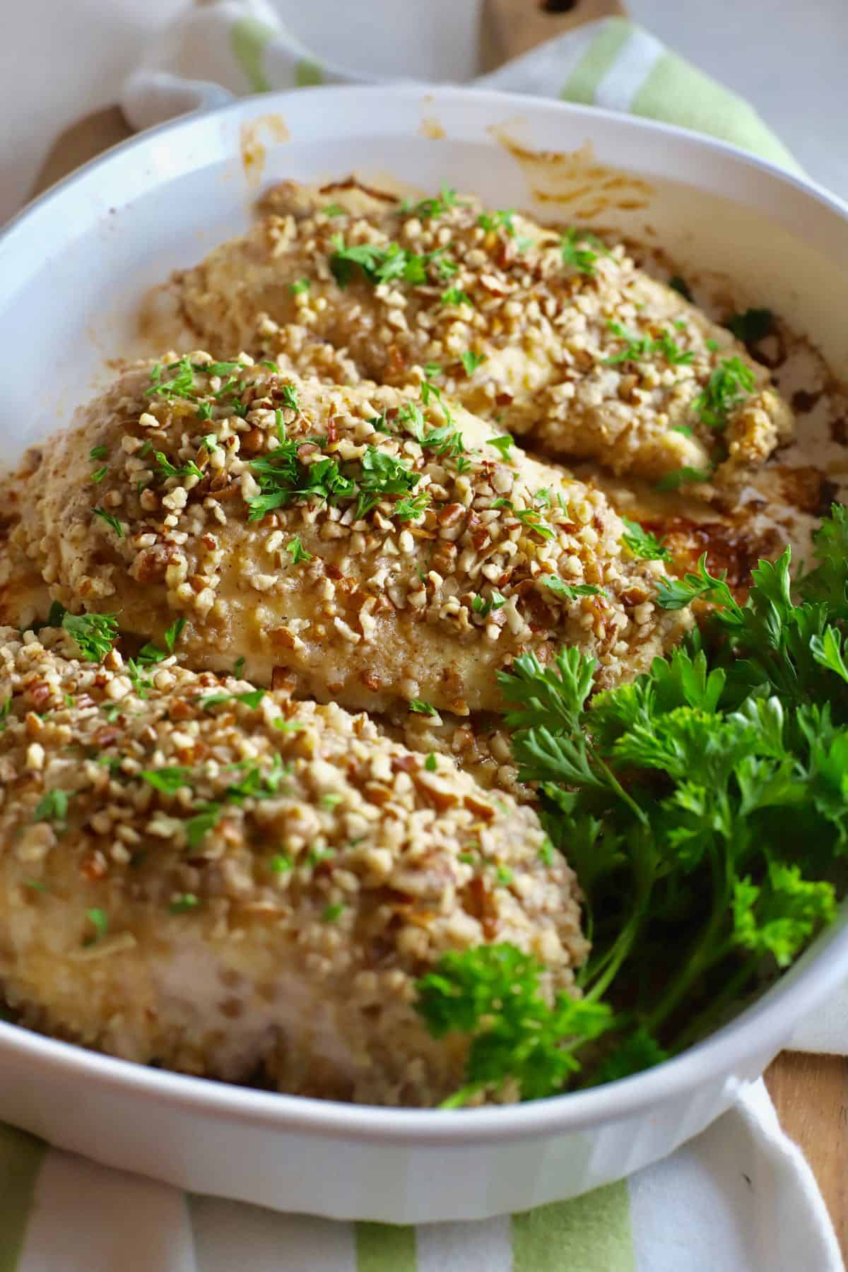 Three chicken breasts in a white baking dish topped with chopped pecans and parsley. 