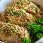 Three pecan crusted chicken breasts in a white baking dish topped with parsley.