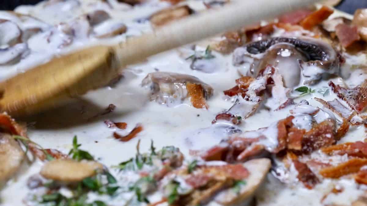 Heavy cream mixed with bacon, mushrooms and thyme in a skillet.