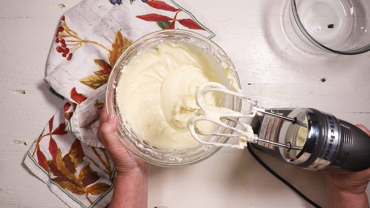 Cream cheese icing in a clear glass bowl next to an electric mixer. 