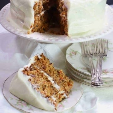 A carrot cake on a cake stand with a slice cut out that is on a plate.