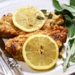 Two pieces of chicken on a white plate garnished with sage and lemon slices.