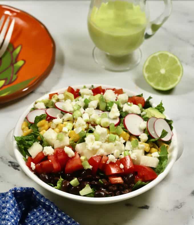 Mexican chopped salad in a white bowl with a small pitcher of honey lime dressing. 