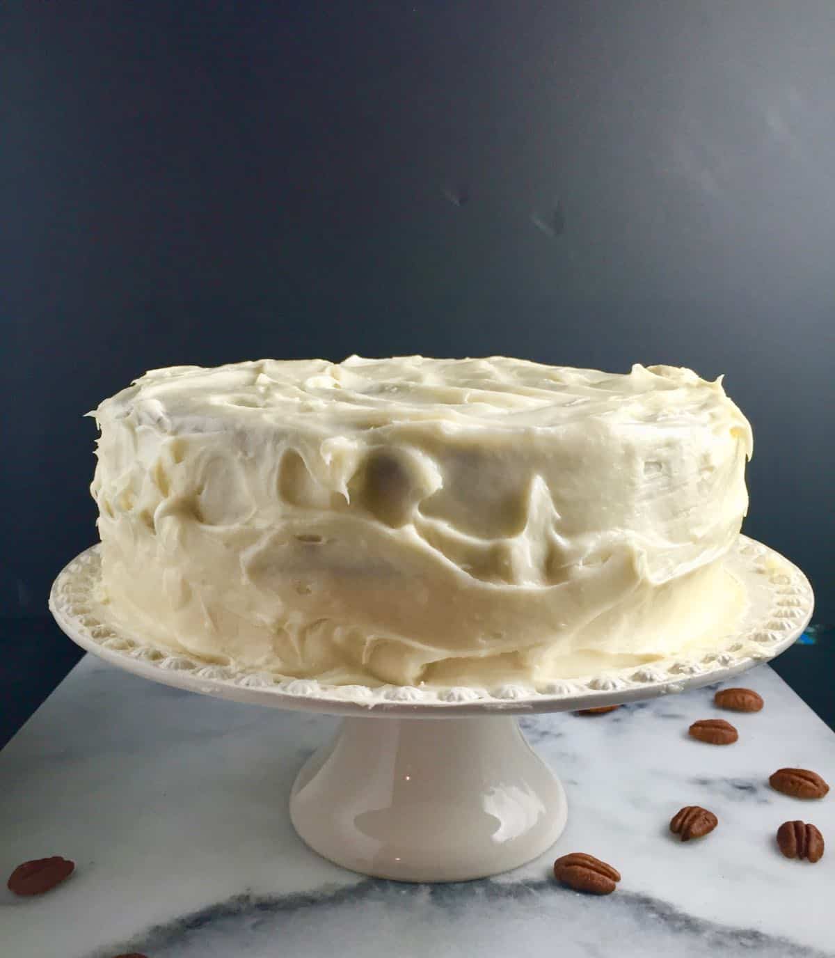 Carrot cake on a white cake platter with pecans on the counter. 