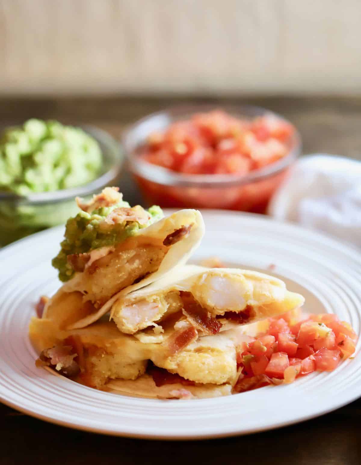 Two quesadillas on a white plate with bowls of chopped tomatoes and guacamole behind them. 