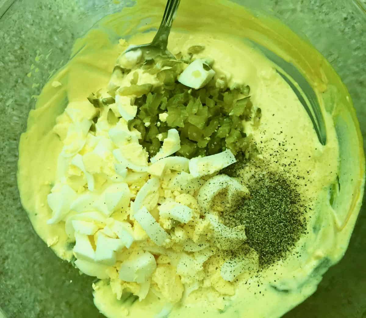 Mayonnaise, mustard, dill pickles and chopped up hard-boiled eggs in a bowl.