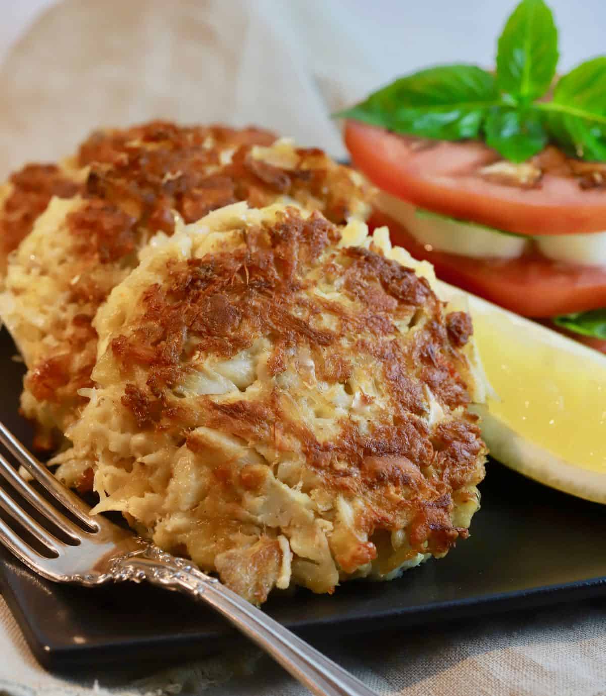 Three crab cakes on a black plate with sliced tomatoes.