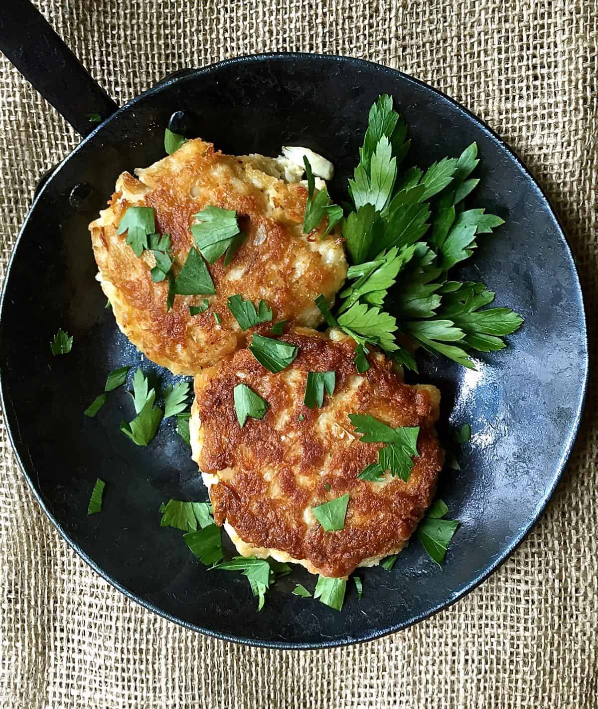 Two golden brown blue crab cakes topped with parsley in a small cast iron skillet.