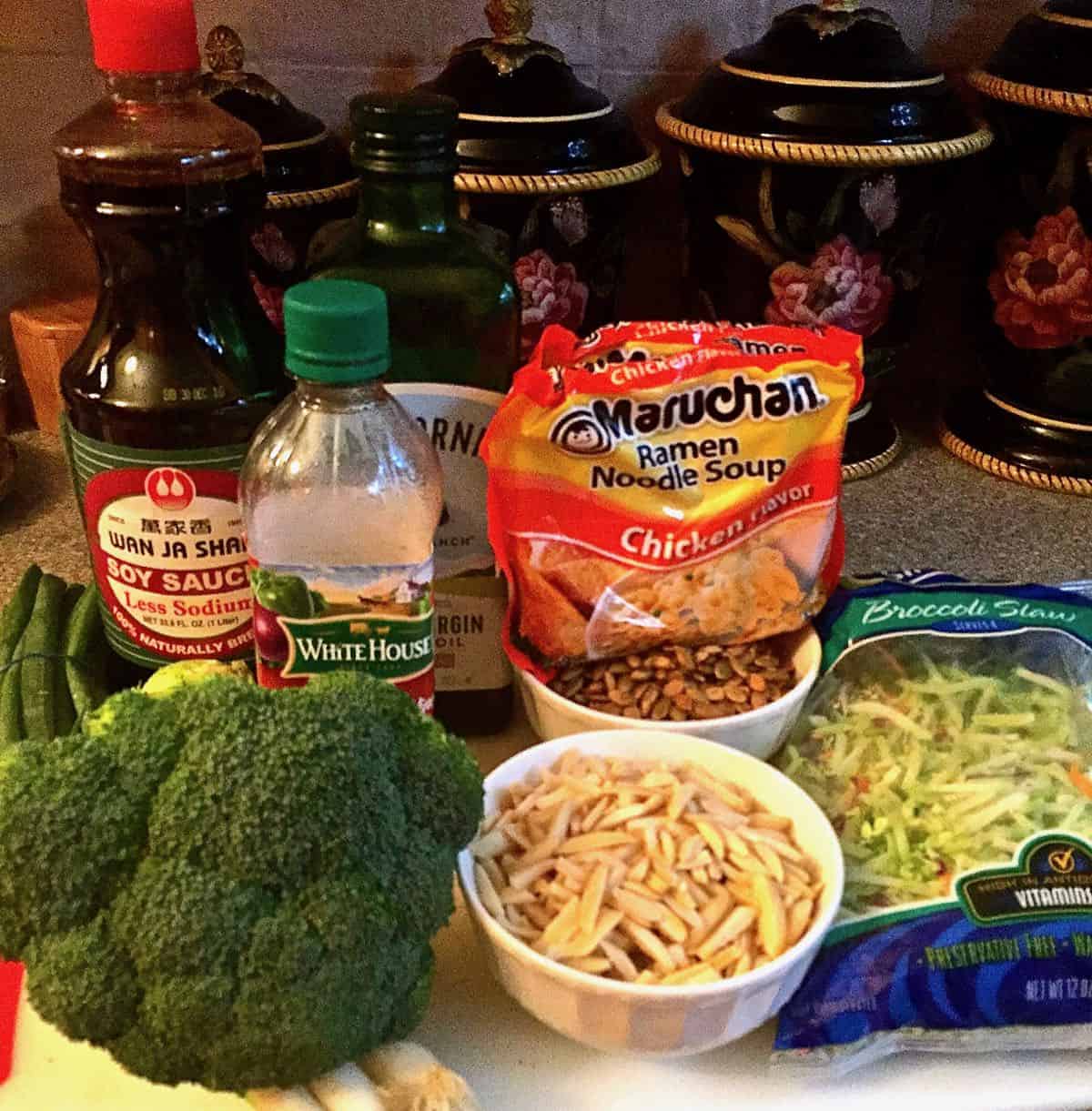 A head of broccoli, slivered almonds, ramen noodles and soy sauce on a counter. 