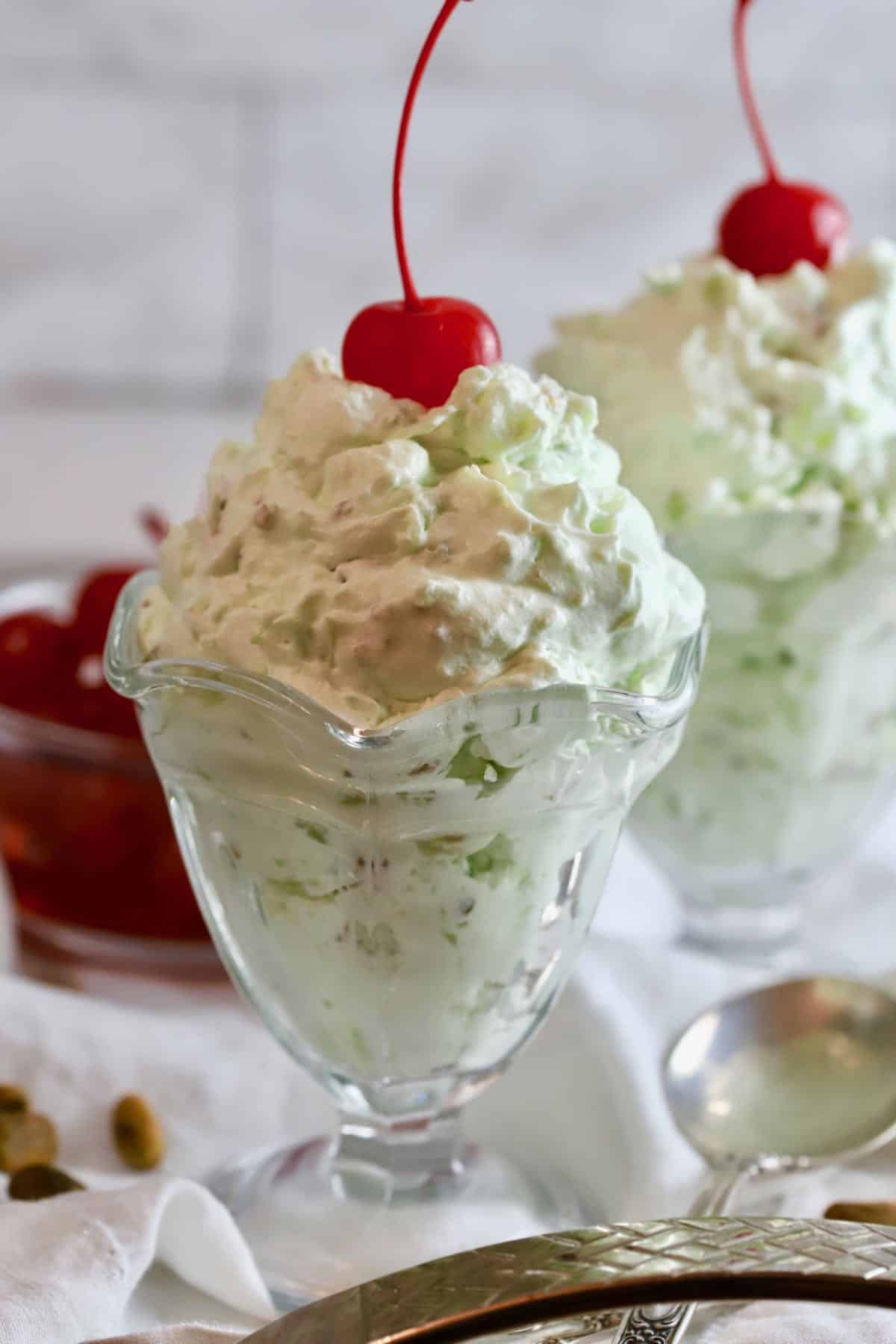 Two glass dessert dishes full of Pistachio Pudding topped with a cherry.