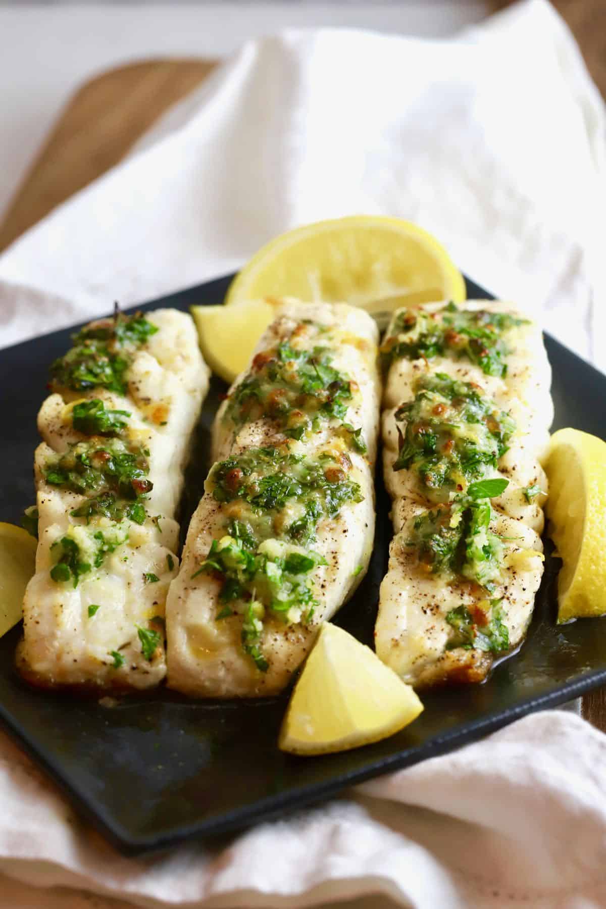 Fish fillets on a black plate garnished with lemon slices. 