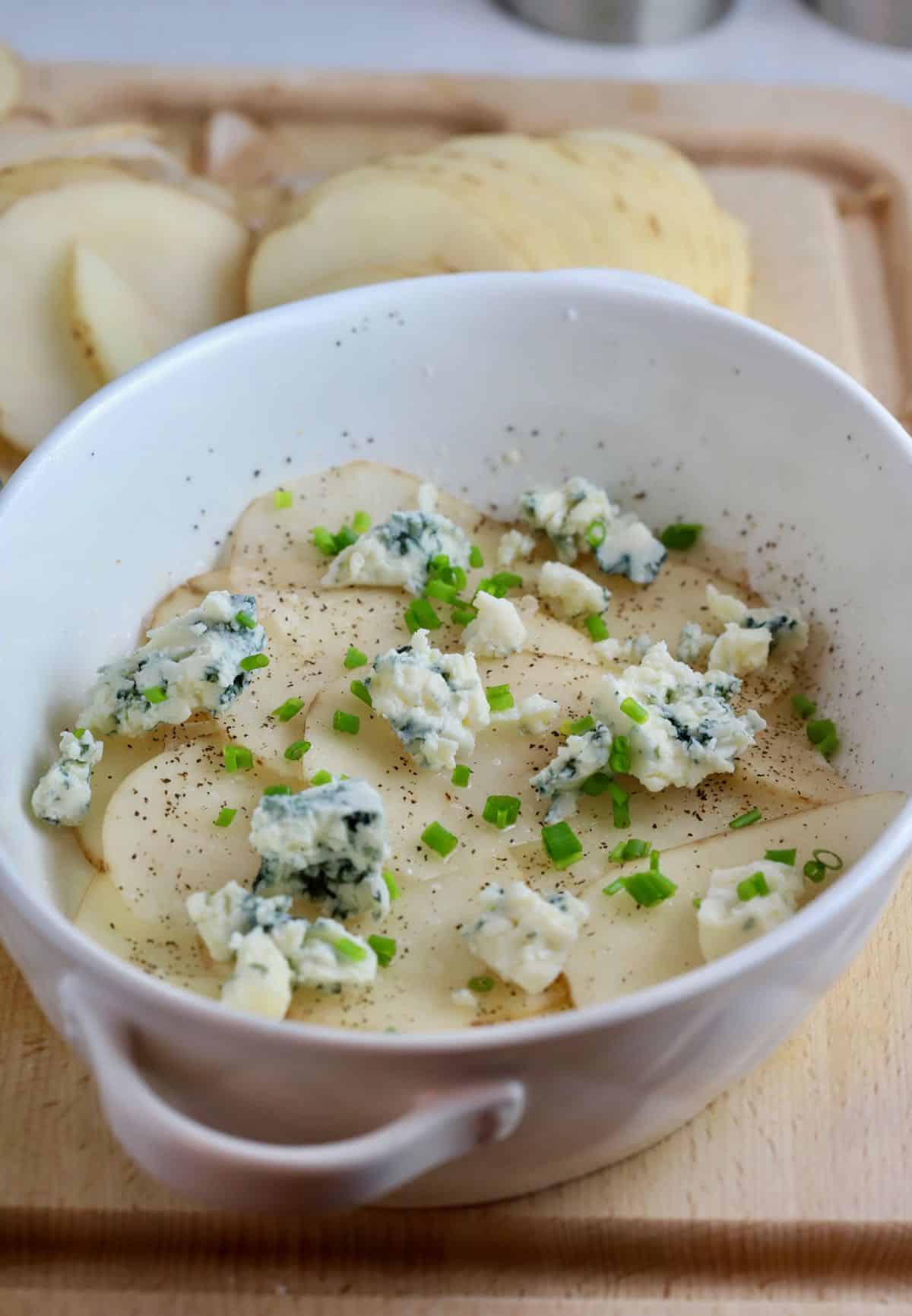 A white baking dish with potato slices topped with chives and blue cheese. 
