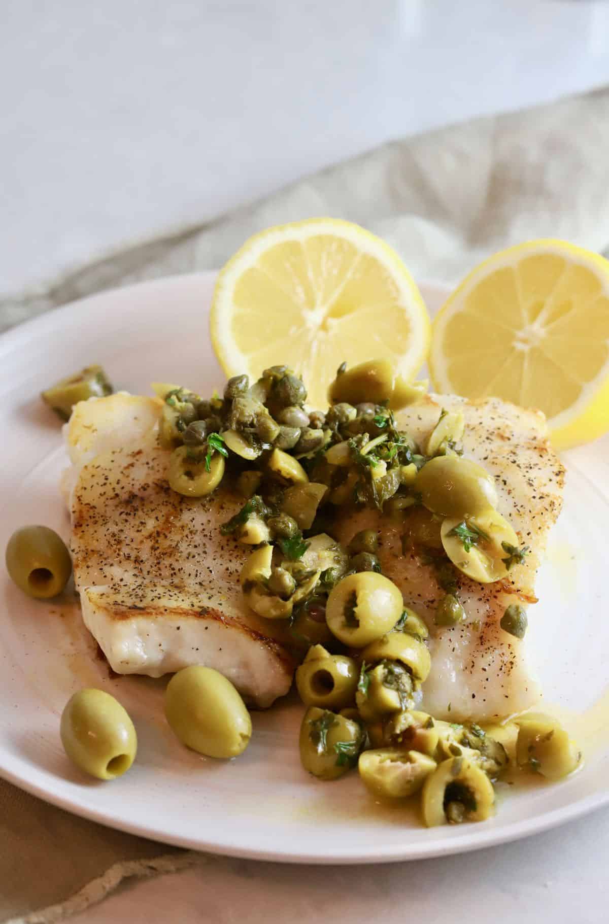Mediterranean grouper on a white plate topped with olives and capers. 