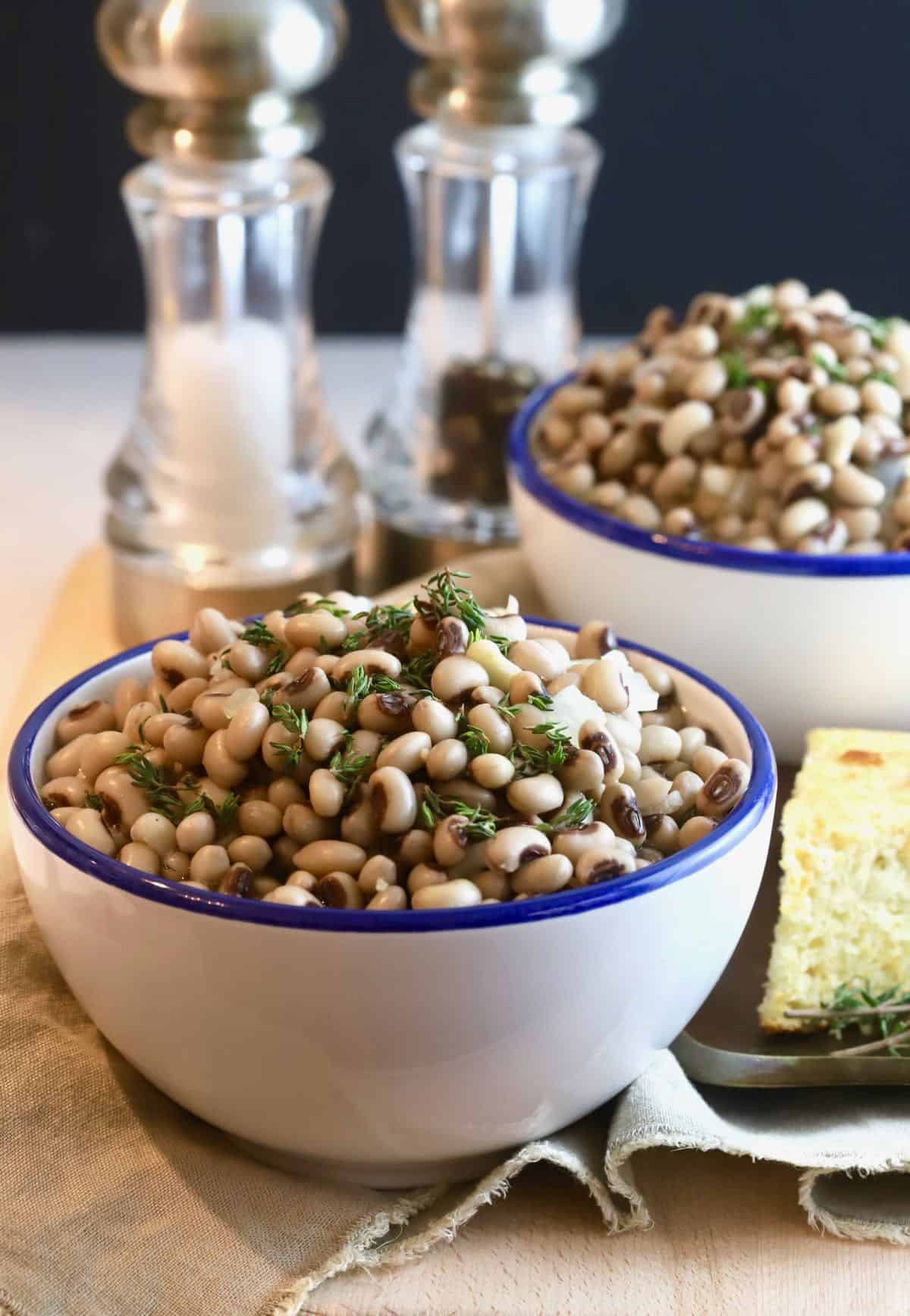 Two bowls of black-eyed peas with salt and pepper shakers in the rear and a slice of cornbread.