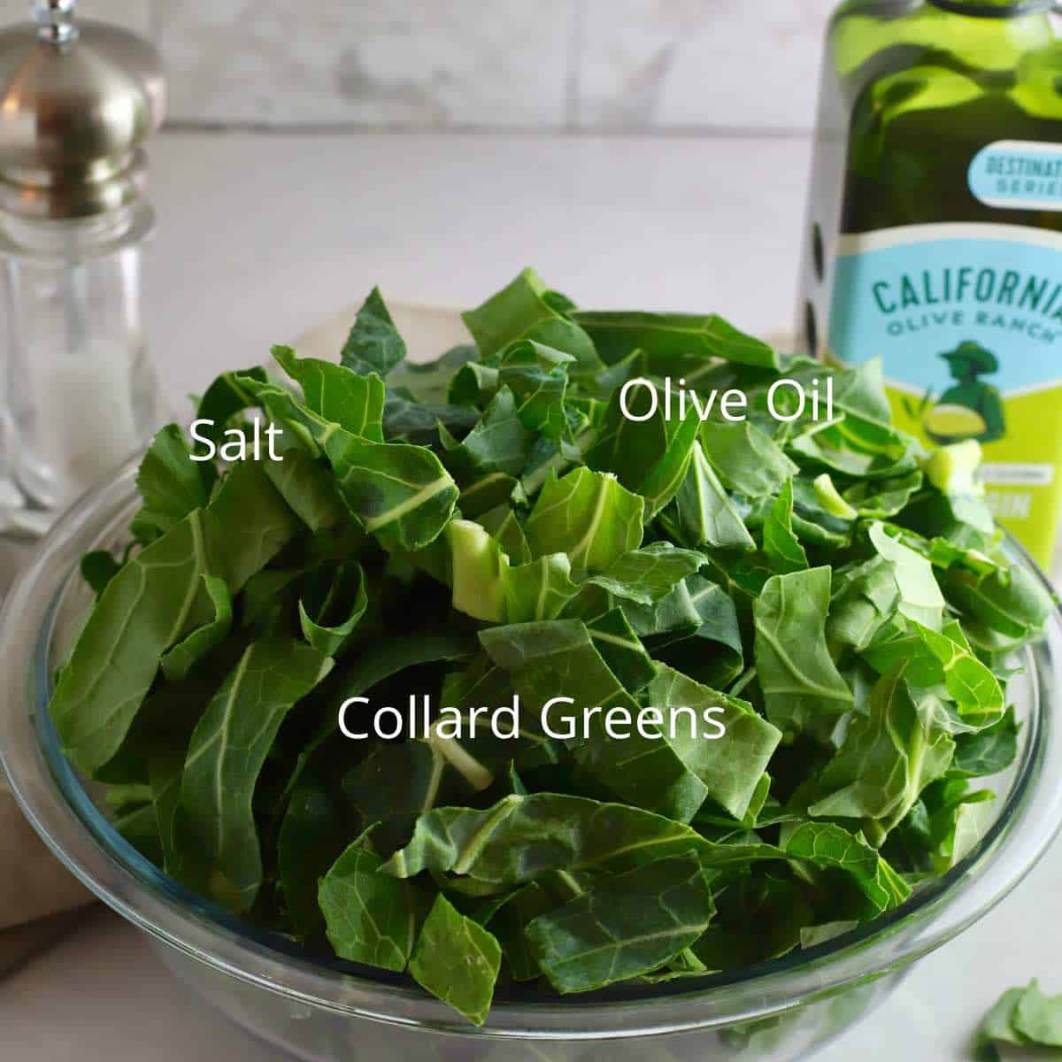 A large bowl of uncooked collard greens, a bottle of olive oil and salt. 