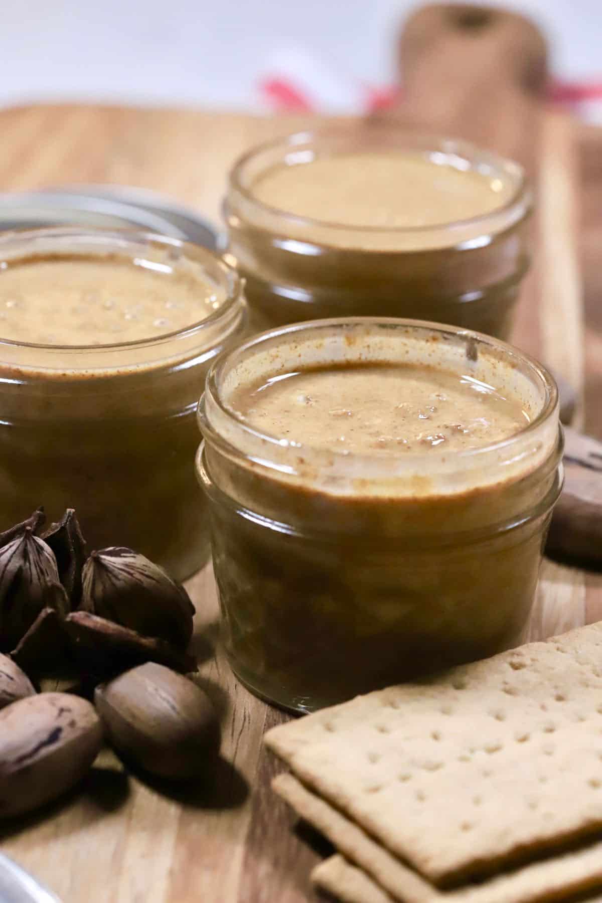 Three glass jars of homemade pecan butter with graham crackers on a cutting board. 
