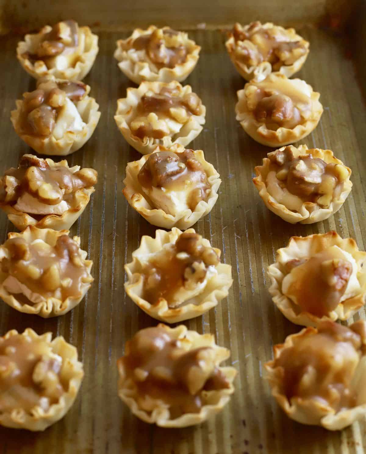 Phyllo shells on a baking sheet full of brie cheese and topped with sugar and pecans. 