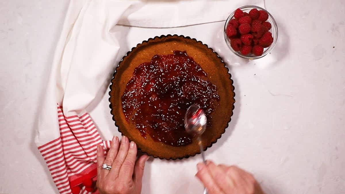 Using a spoon to spread raspberry preserves over a tart crust. 