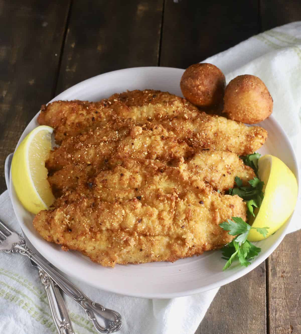 Fried fish fillets on a white platter with hushpuppies and slices of lemon. 