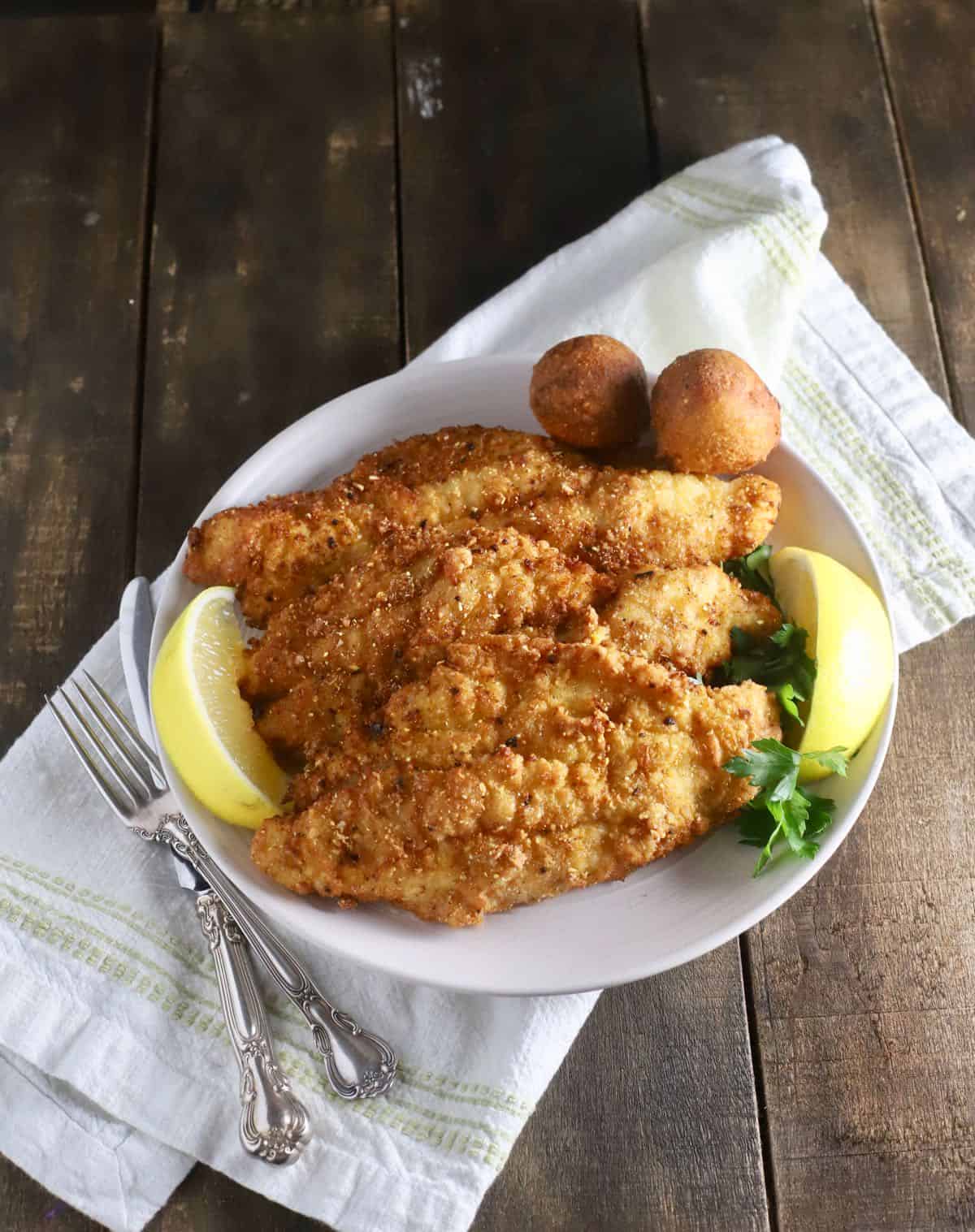 Three fried catfish fillets on a white plate with hushpuppies. 